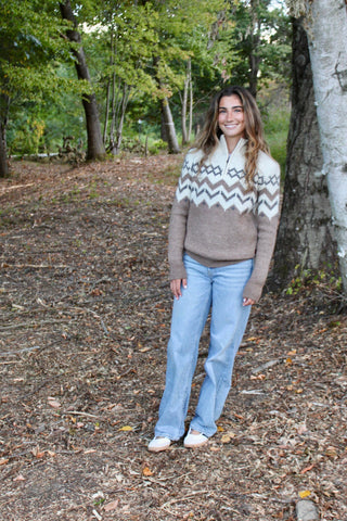 Woman standing in a forest wearing a patterned sweater and jeans.